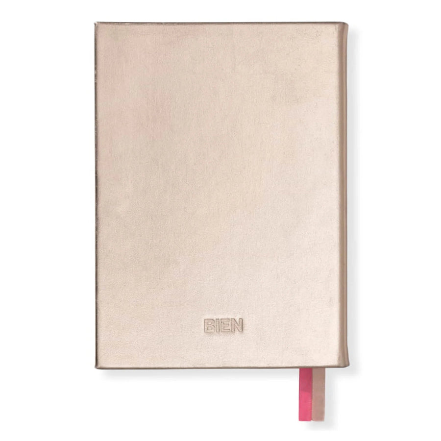 A slim hardcover notebook in a metallic rose gold finish with the brand name 'BIEN' debossed on the lower front and a pink ribbon bookmark peeking out from the bottom right, set against a plain white background.