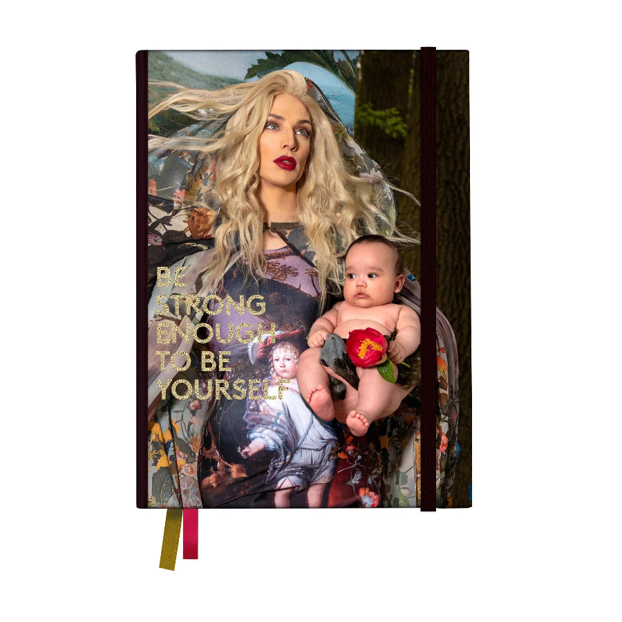 A hardcover journal featuring a surreal collage-style cover art of a blonde woman holding a baby with a red rose, with the text 'BE STRONG ENOUGH TO BE YOURSELF' in gold. The notebook has a black elastic closure and ribbon bookmarks in red and gold, shown on a plain white background.