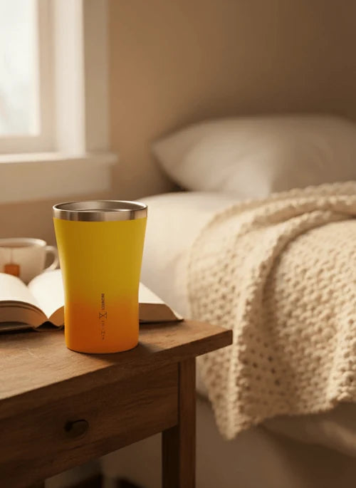 A yellow to orange gradient insulated tumbler with a stainless steel rim sits on a wooden bedside table in a softly lit bedroom setting.