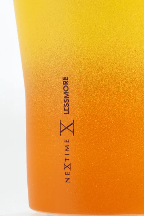 Close-up of a frosted glass tumbler with a yellow-to-orange gradient finish, featuring the vertical black text 'NEXTIME X LESSMORE' on the side.