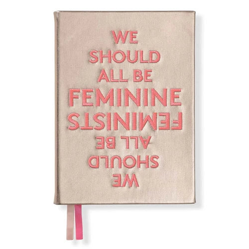 A beige hardcover journal with pink embroidered text reading 'WE SHOULD ALL BE FEMINIST' and a pink ribbon bookmark, shown on a white background.