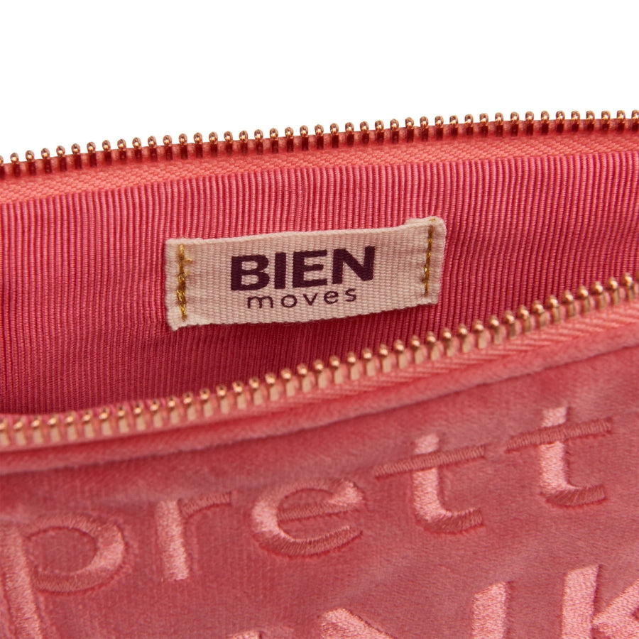 Close-up of a pink zippered pouch with a beige fabric label reading 'BIEN moves' and a rose gold zipper, on a white background.
