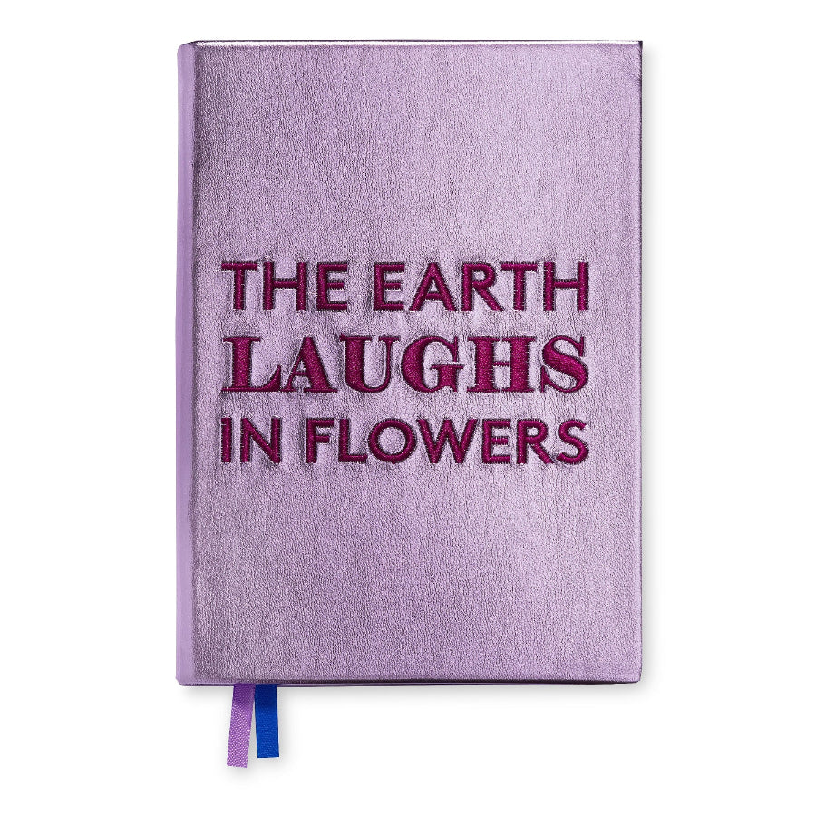 A lavender purple hardcover journal with the phrase 'THE EARTH LAUGHS IN FLOWERS' embossed in dark magenta lettering on the front cover. A blue and purple ribbon bookmark is visible at the bottom edge, set against a plain white background.