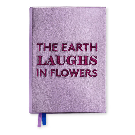 A lavender purple hardcover journal with the phrase 'THE EARTH LAUGHS IN FLOWERS' embossed in dark magenta lettering on the front cover. A blue and purple ribbon bookmark is visible at the bottom edge, set against a plain white background.