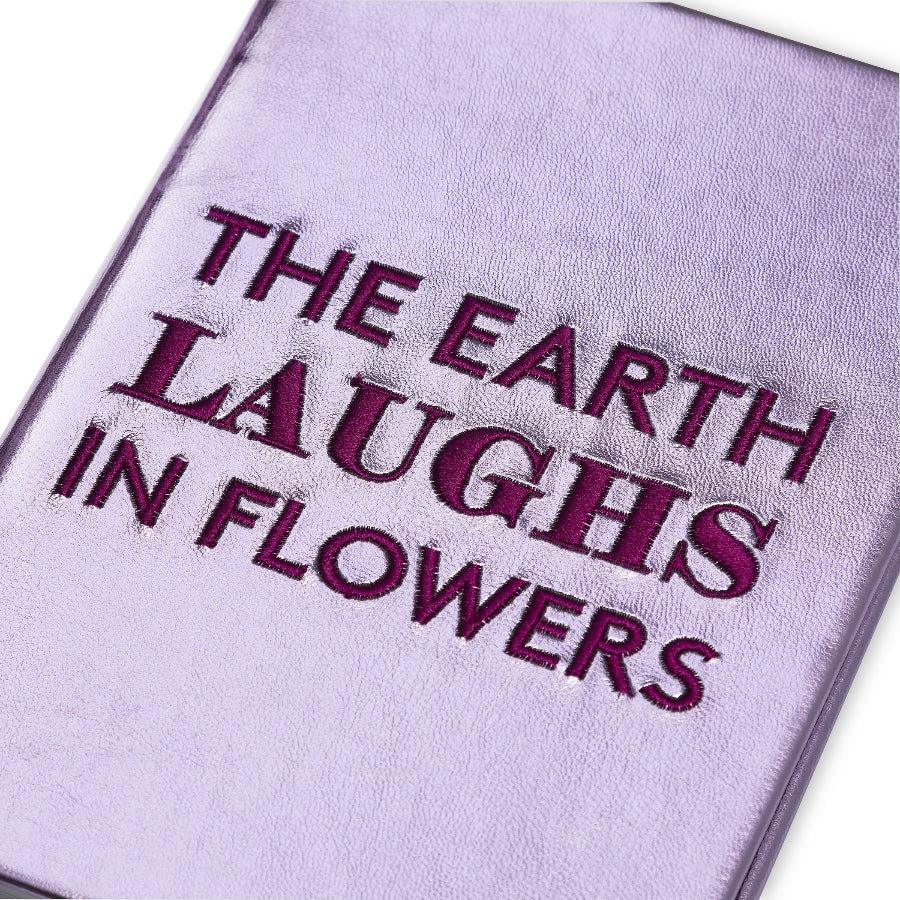 A close-up of a lavender-purple textured book cover with the phrase 'THE EARTH LAUGHS IN FLOWERS' embroidered in dark purple lettering, shown against a white background.