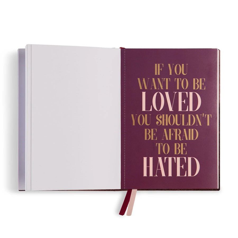 An open hardcover journal or notebook on a white background. The right page is a deep plum purple with metallic gold text that reads, 'IF YOU WANT TO BE LOVED YOU SHOULDN'T BE AFRAID TO BE HATED.