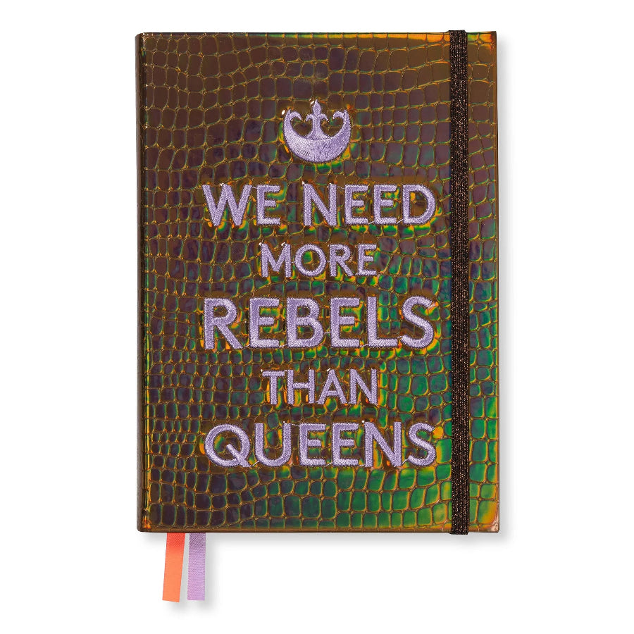 A hardcover notebook with a textured, iridescent reptile skin cover. The cover features a purple glitter crown graphic above the text 'WE NEED MORE REBELS THAN QUEENS' in glittery purple lettering.